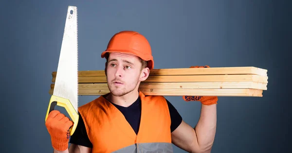 Holzhandwerkliches Konzept. Mann, Handwerker mit Helm, harter Hut, Handsäge, Blick auf scharfe Klinge, grauer Hintergrund. Tischler, Holzarbeiter, Arbeiter, Bauarbeiter mit nachdenklichem Gesicht trägt Holzbalken auf der Schulter — Stockfoto