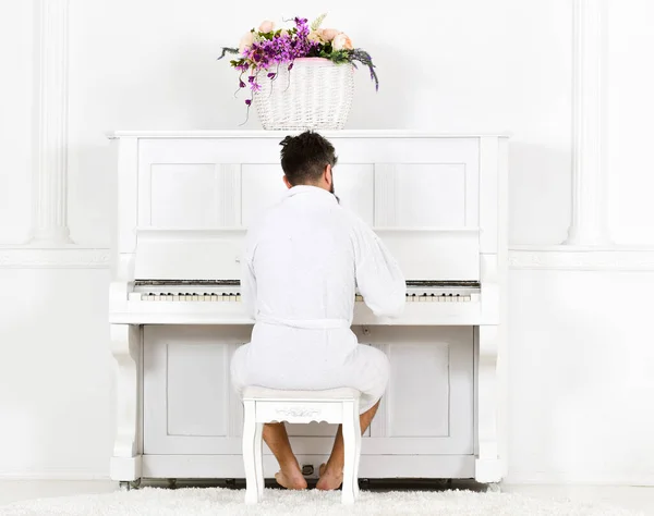 バスローブの男はピアノを演奏しながら朝を楽しむ。バスローブの男眠い背景に白いインテリアでピアノ楽器の前に座って、リアビュー。才能ある音楽家のコンセプト — ストック写真