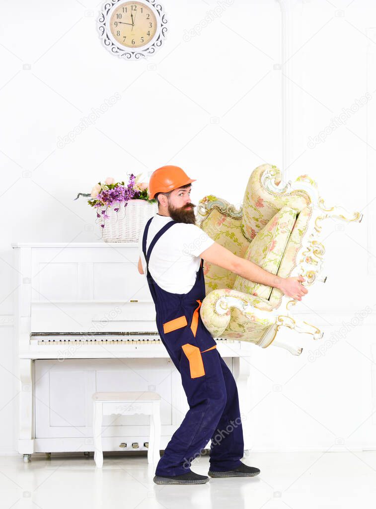 Worker relocating vintage furniture in white room. Careful mover carrying antique armchair in front of piano