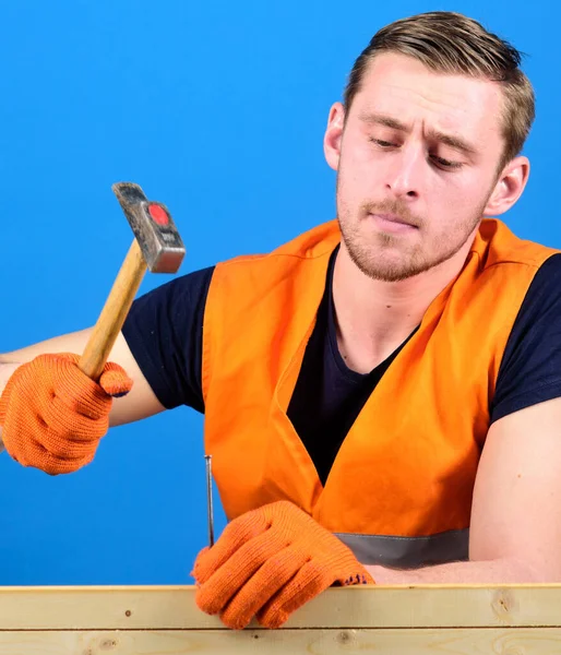 Building and construction concept. Carpenter, woodworker on busy face hammering nail into wooden board. Man, labourer, handyman in bright vest and protective gloves handcrafting, blue background