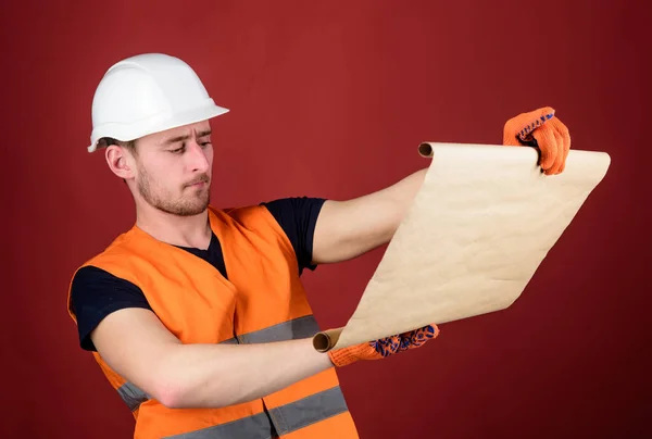 Foreman concept. Engineer, architect, builder on strict face holds blueprint in hands, supervises construction site. Male contractor with house plans wearing hard hat, controls works, red background. — Stock Photo, Image