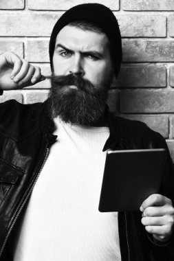 Sakallı, beyaz tenli hippi bej tuğla duvar stüdyosunun arka planında tablet taşıyor.