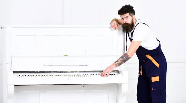 Homem com barba e bigode, trabalhador de macacão inclinado para piano, fundo branco. Carregador move instrumento de piano. Courier entrega móveis em caso de mudança, realocação. Conceito de serviço de entrega — Fotografia de Stock