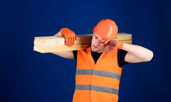 Carpenter, woodworker, labourer, builder on tired face carries wooden beam on shoulder. Man in helmet and protective gloves wiping sweat from forehead, blue background. Tired labourer concept. — Stock Photo, Image