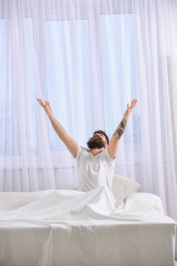 Gülümseyen adam kollarını yukarı kaldırıyor. Dinlenme konsepti. Gömlekli adam yatakta oturur, arka planda beyaz perdeler. Sakallı ve bıyıklı maço. Esniyor, dinleniyor.