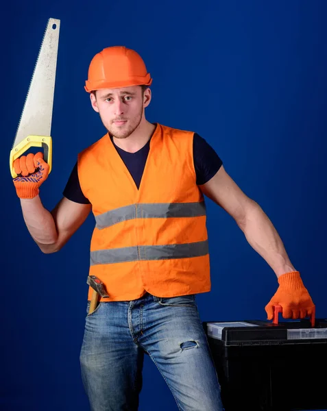 Man in helm, harde hoed draagt gereedschapskist en houdt handzaag, blauwe achtergrond. Werknemer, reparateur, reparateur op ernstig gezicht draagt gereedschapskist, klaar voor reparatie. Een klusjesman concept. Timmerman met handzaag — Stockfoto