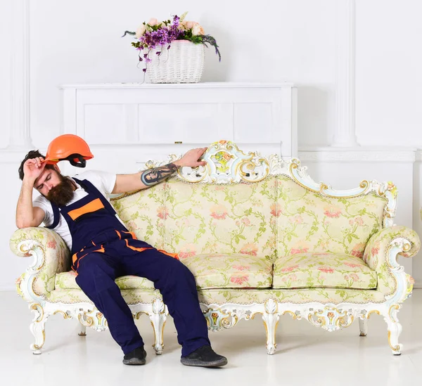 Loader sit on sofa, having rest. Man with beard, worker in overalls and helmet sits on couch tired, white background. Exhausted loader concept. Courier relaxing while moving furniture, relocation.