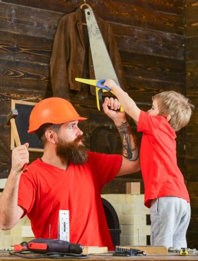 Babalık kavramı. Neşeli çocuk elinde oyuncak testereyle babasıyla el işi yaparken eğleniyor. Baba, koruyucu kasklı ebeveyn oğluna okulda farklı aletler kullanmayı öğretiyor..