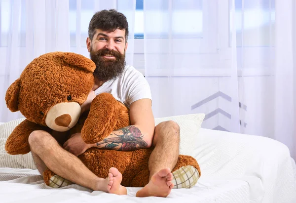 Kerl mit fröhlichem Gesicht umarmt riesigen Teddybär. Infantilismus-Konzept. Macho mit Bart und Schnurrbart entspannt mit Plüschtier nach dem Mittagsschlaf, Ruhe. Mann sitzt auf Bett und umarmt großes Spielzeug, weiße Vorhänge vor Hintergrund — Stockfoto