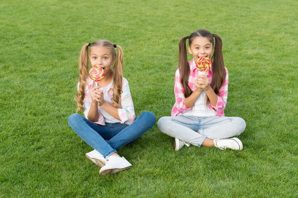 Sciogliti in bocca il dolce. I bambini felici mangiano il dessert di caramella su bastoni. Le bambine si siedono sull'erba verde. Mi piace il dolce dessert. Negozio di caramelle. Dolciumi a base di zucchero. Vacanze estive. Tempo del dolce. — Foto Stock