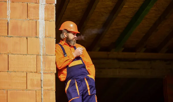 Építőmunkás cigarettázik. Az épület építés alatt áll. Szakállas férfi egyenruhában és keménykalapban. Másolás. A szerelő mérnök sisakot visel a helyszínen. Fáradt építész pihenni. pihenés ideje — Stock Fotó