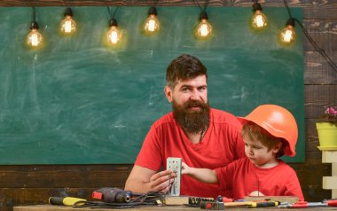 Sakallı baba küçük oğluna sınıfta alet kullanmayı öğretiyor. Arka planda karatahta var. Küçük asistan konsepti. Çocuk, koruyucu kasklı çocuk, neşeli babasıyla tamir ediyor..