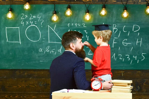 Skolpauskonceptet. Lärare med skägg, far undervisar lille son i klassrummet, tavlan på bakgrunden. Pojke, barn i examen mössa leka med pappa, ha kul och koppla av under skollov. — Stockfoto