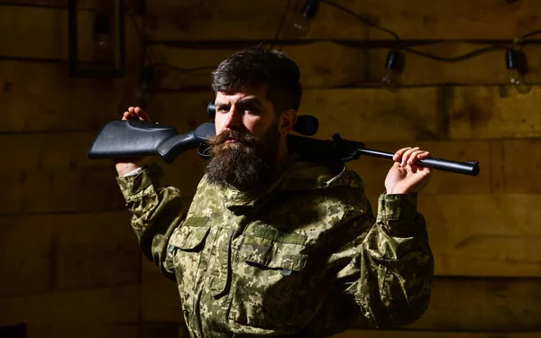 Masculinity concept. Man, gamekeeper with beard wears camouflage clothing, carries rifle on shoulders, wooden interior background. Hunter, brutal hipster on strict face with gun ready for hunting. — Stock Photo, Image