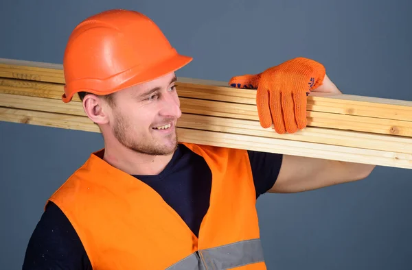 头戴头盔、头戴硬礼帽、戴着防护手套的男人拿着木梁,灰色背景.勤劳的概念。木匠，木匠，工人，面带微笑的建筑工人，肩上扛着木梁. — 图库照片