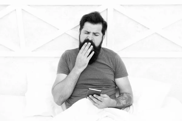 Le lleva mucho tiempo despertarse. Hombre somnoliento. El barbudo usa el teléfono móvil en la cama. El hombre bosteza por la mañana. Hombre sin afeitar con barba y pelo de bigote. Estilo de vida móvil. Despertando. Buenos días. —  Fotos de Stock