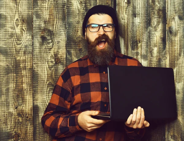 Bearded brutal caucasian hipster holding laptop on brown vintage wooden studio background — Stock Photo, Image