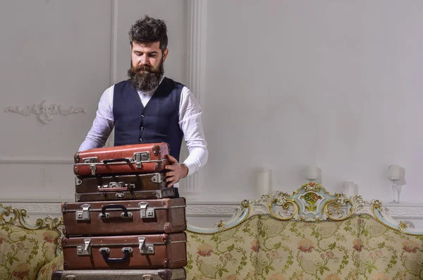 Hombre, viajero con barba y bigote equipaje de embalaje antes del viaje, fondo interior de lujo. Macho elegante en la cara reflexiva de pie cerca de pila de maleta vintage. Equipaje y concepto de viaje . — Foto de Stock