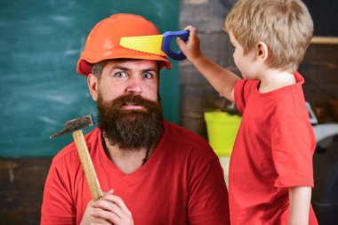 Oyuncak testereyle oynayan neşeli bir çocuk, babayla alet kullanmayı öğreniyor. Babalık kavramı. Baba, koruyucu başlıklı sakallı bir ebeveyn küçük oğluna okulda farklı aletler kullanmayı öğretiyor..