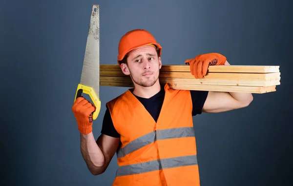 Menuisier, menuisier, ouvrier, constructeur sur un visage réfléchi porte des poutres en bois sur l'épaule. Homme, bricoleur au casque, casque dur tient scie à main et poutres en bois, fond gris. Concept d'artisanat bois . — Photo