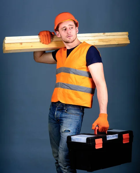 Carpinteiro, marceneiro, trabalhador, construtor no rosto calmo carrega vigas de madeira no ombro. Conceito de construção e marcenaria. Homem de capacete, chapéu duro segura caixa de ferramentas e vigas de madeira, fundo cinza . — Fotografia de Stock