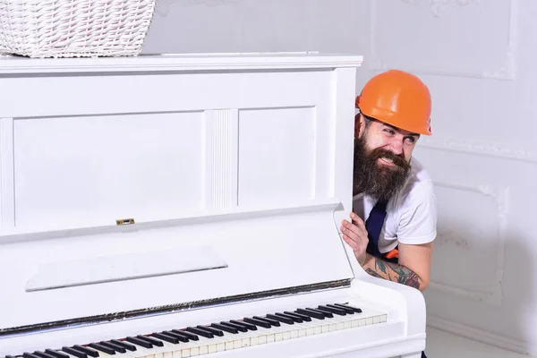 Heavy loads concept. Loader moves piano instrument. Man with beard worker in helmet and overalls pushes, put efforts to move piano, white background. Courier delivers furniture, move out, relocation