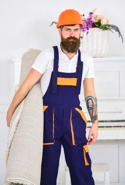 Cleaning service concept. Courier delivers furniture in case of move out, relocation. Loader wrapped carpet into roll. Man with beard, worker in overalls and helmet carries carpet, white background.