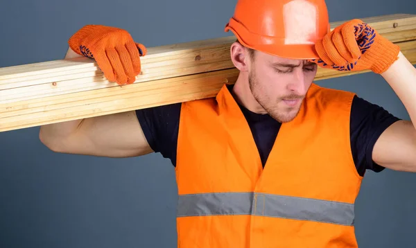 Ξυλουργός, ξυλουργός, δυνατός χτίστης στο πολυάσχολο πρόσωπο κουβαλάει ξύλινη δοκό στον ώμο. Ο άνθρωπος με προστατευτικά γάντια κρατά το προστατευτικό κάλυμμα του σκληρού καπέλου, γκρι φόντο. Έννοια ασφάλειας και προστασίας. — Φωτογραφία Αρχείου