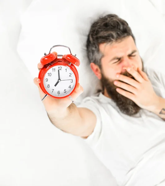 El hombre con la cara bostezante yace en la almohada, sostiene el despertador . —  Fotos de Stock