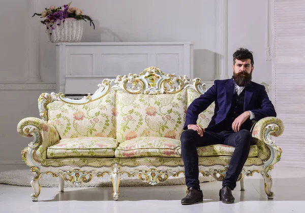Mann mit Bart und Schnurrbart im modischen klassischen Anzug sitzt auf altmodischem Sofa oder Sofa. Macho attraktiv und elegant mit ernstem Gesicht und nachdenklichem Ausdruck. Mode- und Stilkonzept — Stockfoto