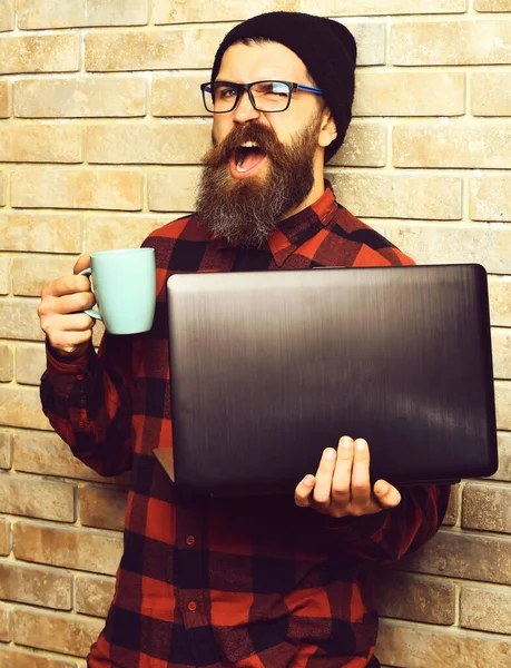 Bearded brutal caucasian hipster holding laptop with cup — Stock Photo, Image
