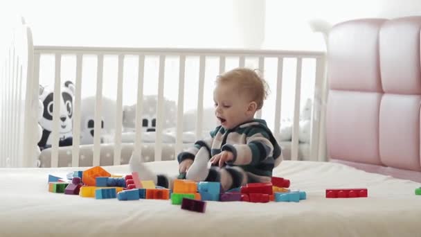 Portret rocznego chłopca bawiącego się kolorowymi plastikowymi klockami lego, siedzącego na łóżku. Maluch dobrze się bawi i buduje z cegieł konstrukcyjnych. Wczesna nauka. Zabawki rozwojowe — Wideo stockowe