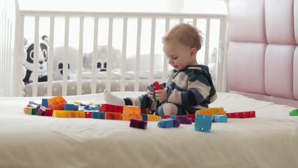Peuter plezier hebben en bouwen van bouwstenen. Vroeg leren. Ontwikkelspeelgoed. Portret van een jaar oude baby jongen die speelt met kleurrijke plastic bakstenen lego, zittend op het bed. — Stockvideo
