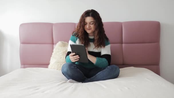 Mujer bonita y joven en su cama usando su tableta, navega por la red social . — Vídeos de Stock