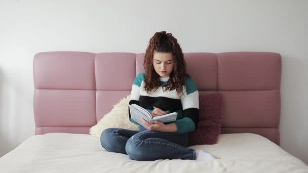 En vacker ung kvinna på sängen som skriver dagbok. Leende brunett dam med nätbok och penna i händerna. — Stockvideo