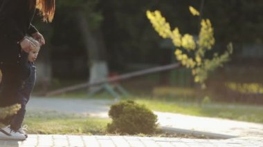Bebeğin annesiyle ilk adımı atması parkta işe yarıyor. Küçük şirin çocuk annesinin elini tutuyor ve kaldırımda ilk adımlarını atıyor. Açık havada..