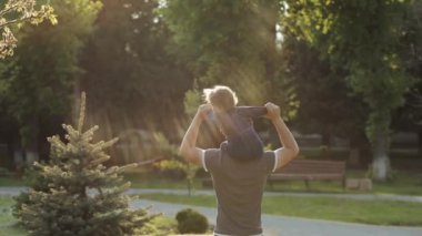 Baba ve Oğul Sunset 'te parkta yürüyor. İki Yaşındaki Çocuk Ebeveynlerin Omuzlarında Oturuyor. Mutlu Aile Kavramı.