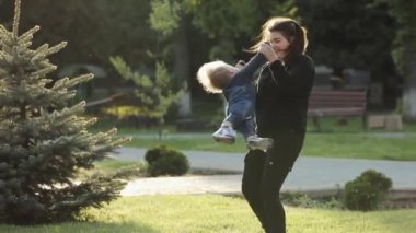 Kadın, sevimli bebeğini kaldırır ve gülümseyerek onu döndürmeye başlar. Mutlu an ebeveyn ve anne, oğluyla parkta oynayarak vakit geçirir. .