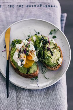 Mikro yeşil ve haşlanmış yumurtalı avokado tostu. Yumurta sarısı tabağa dökülüyor. Minimalist yemek tasarımı. Gri arka plan. Avokado ekmeği dilimleri. Mikro yeşiller. Sağlıklı kahvaltı..