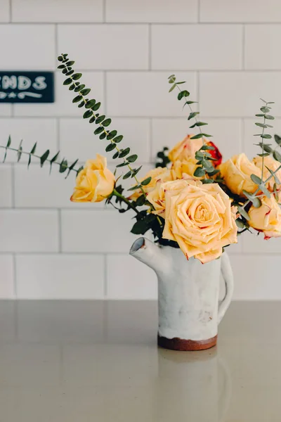 Yellow large roses and eucalyptus branches in the ceramic craft white vase. Kitchen counter top. Lifestyle bright kitchen. Coffee space in the kitchen. Grey and white colors. Bouquet of yellow roses.