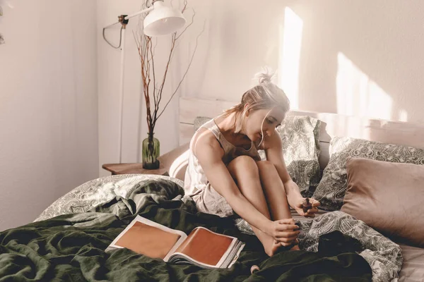 Woman in bed with magazine. Relaxation time. Woman beauty routine. Girly leisure time. Bright bedroom. Do nails. Read magazine. Blonde lady spend her own time. Modern interior design. Linen dress.