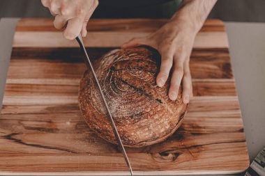 Ev yapımı ekşi ekmek. Buğday çavdar unu. Adam kesme tahtasındaki ekmeği kesiyor. - El yapımı. Gıda trendleri. Tahta kesme tahtası. Erkek eli. Yatay manzara görüntüsü. Evde kal, ekmek pişir..