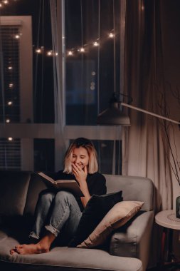 Kız akşamları kanepede kitap okur. Gece lambası. Rahatlatıcı bir akşam. Büyük pencere. Modern oturma odası. Kadife yastıklar. Gece lambası. Sarışın kadın kurgu kitabı okuyor. Karantina evde kalacak..