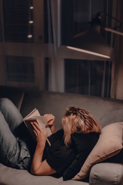 Kız akşamları kanepede kitap okur. Gece lambası. Rahatlatıcı bir akşam. Büyük pencere. Modern oturma odası. Kadife yastıklar. Gece lambası. Sarışın kadın kurgu kitabı okuyor. Karantina evde kalacak..