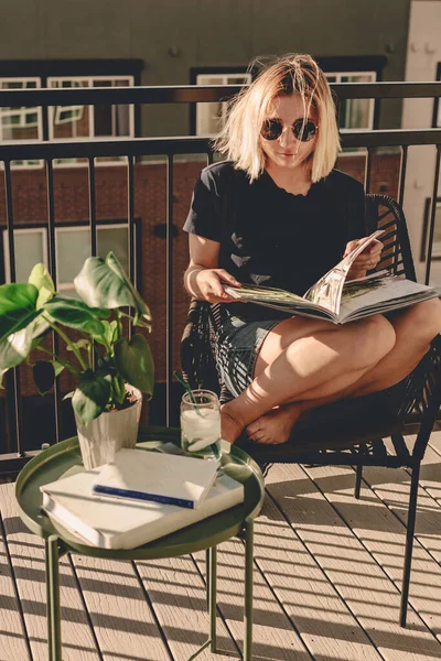 Lady in summer outfit and sunglasses reads book on the balcony. View from the balcony. Sunny day. Relaxing time. Cucumber iced water. Blonde girl stays home. Large balcony. Patio. Warm sunny day.