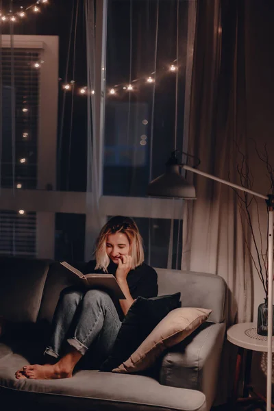 Girl reads book in the evening on couch. Night lamp light. Relaxing evening. Large window. Modern living room. Velvet pillows. Night light. Blonde woman reads fiction book. Quarantine stay at home.