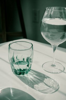 Beyaz masada bir bardak su. Gözlükler ve gölgeler arasında sert bir ışık. Temiz minimalist çağdaş sanat. Kavramsal görüntü. Yeşil ve beyaz temiz renkler. Bardaktaki saf su. Basit görsel kavram.