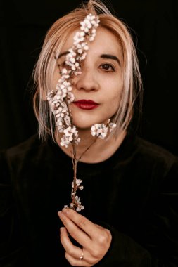 Güzel genç kadın yüzü. Kırmızı seksi dudaklar, güzel gözler. Kameranın önünde sarışın kız portresi. Siyah arka plan. Yüzünde duygular var. Minimalist bir kavram. Makyaj. Güzellik ve moda. Yüzünde çiçekler