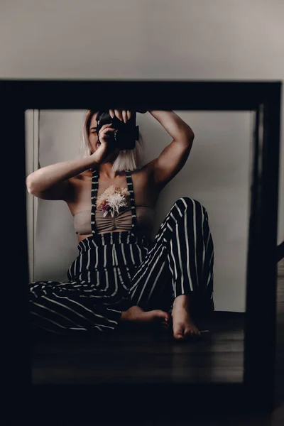 Woman in striped pants sits near window. Self portrait. Mirror reflection. Moody woman photography. Artistic concept. Blonde lady. Sensual, tender, innocence. Square mirror. Black frame. Camera