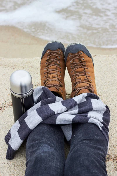 Feet in yellow boots on the background seas. Wrapped in a striped scarf.  With a thermos of hot tea, coffee
